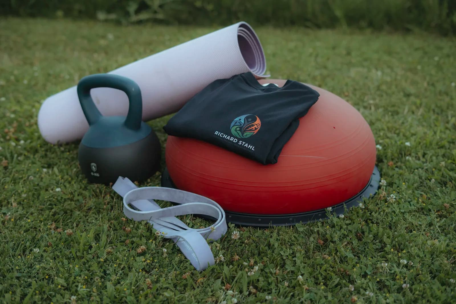 Trainingsequipment von Richard Stahl auf einer Wiese – Kettlebell, Balancetrainer, Matte und Shirt mit Logo